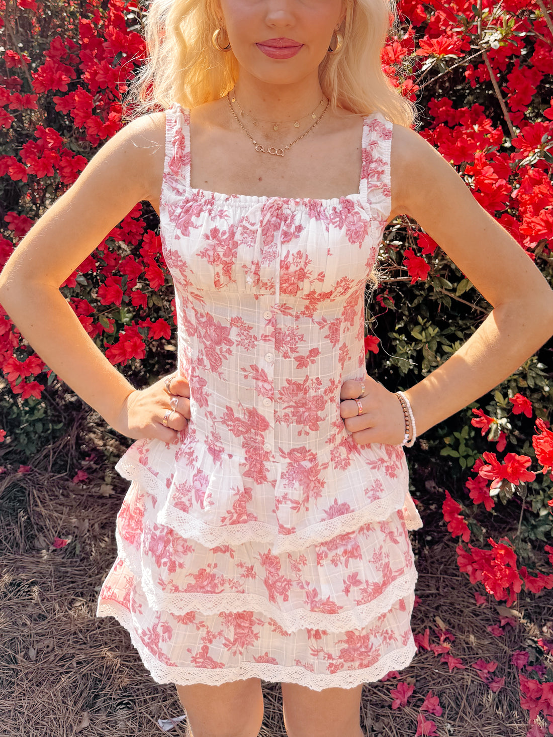 Pink floral mini dress with tiered lace trim, modeled outdoors amidst vibrant red blooms.
