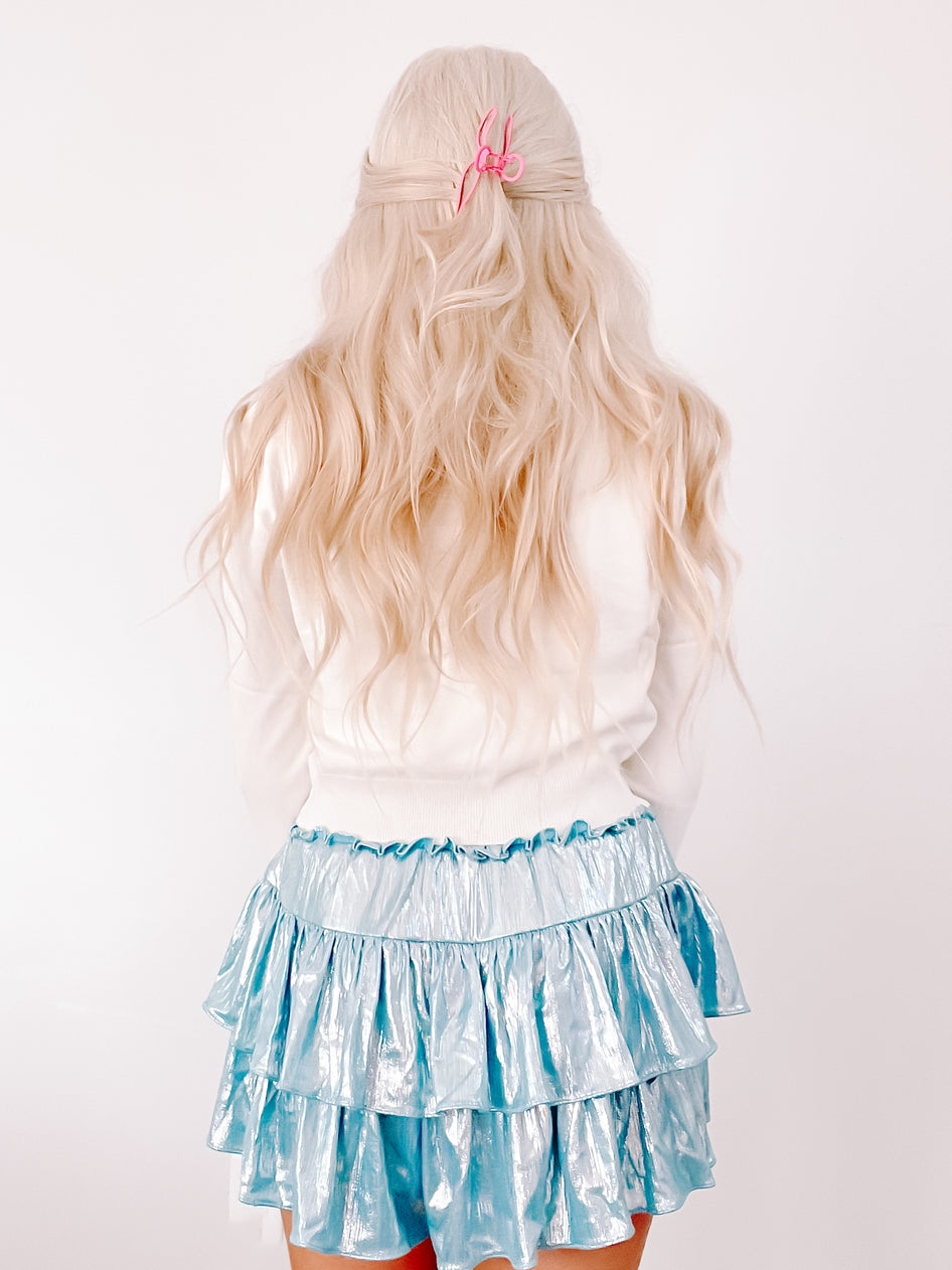 Model wearing a white sweater and a light blue metallic ruffled mini skirt against a white background.
