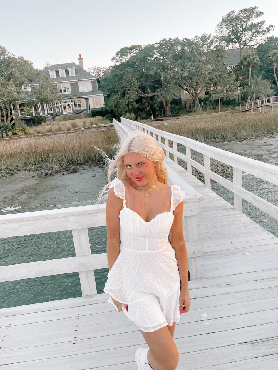 Woman in white eyelet dress on a pier overlooking calm water and houses.

