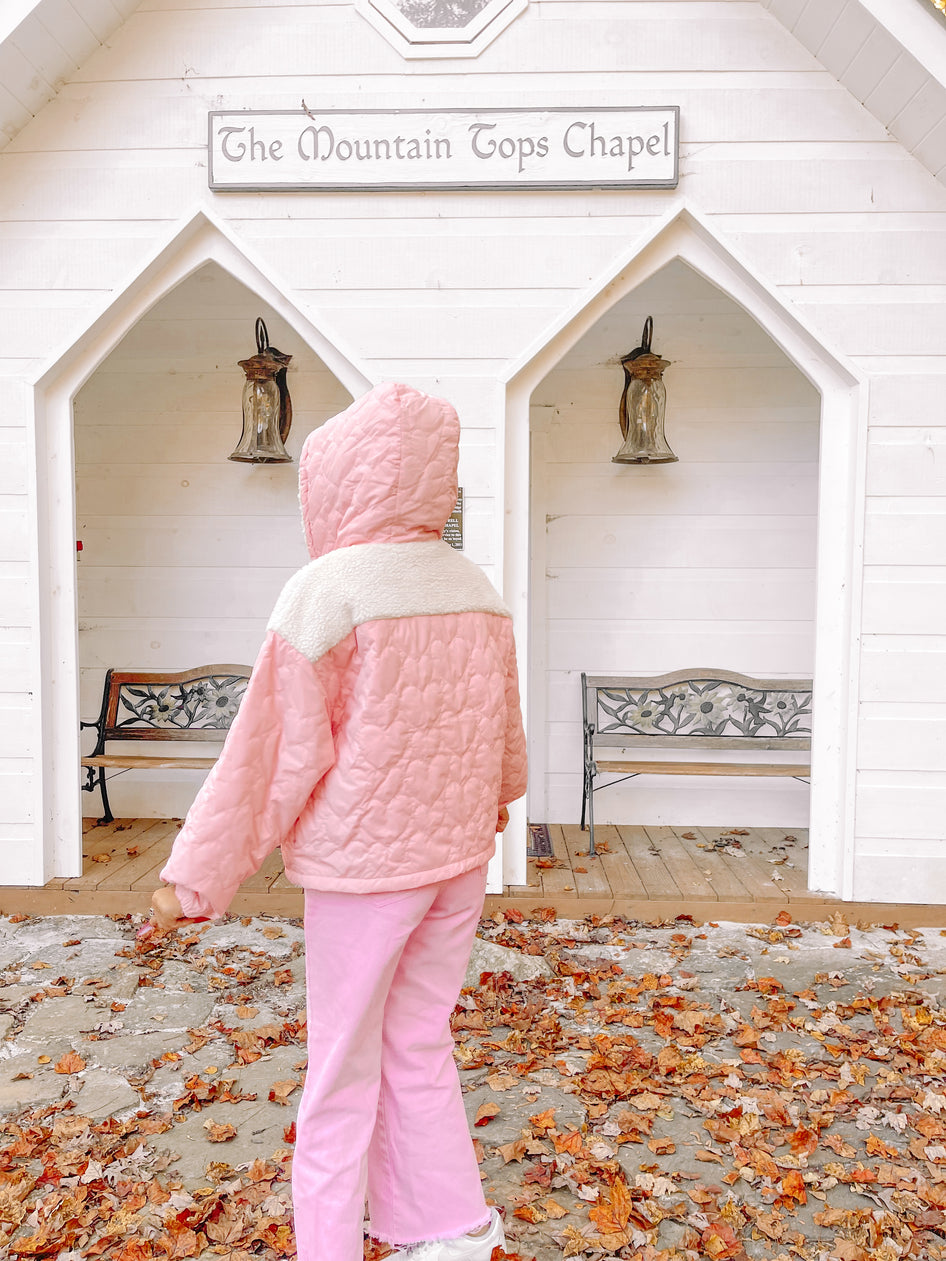 Pink quilted jacket with sherpa lining, worn outdoors near a white chapel in autumn.
