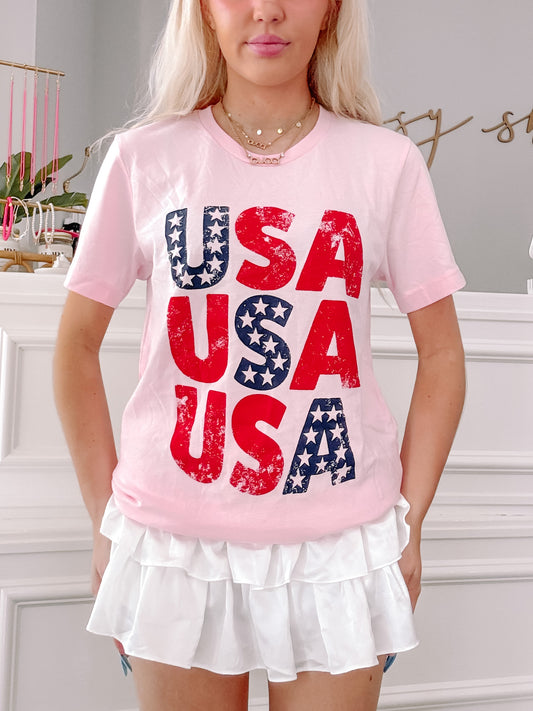 Pink USA graphic tee with distressed red and blue lettering, stars, and a model wearing a white ruffled skirt.