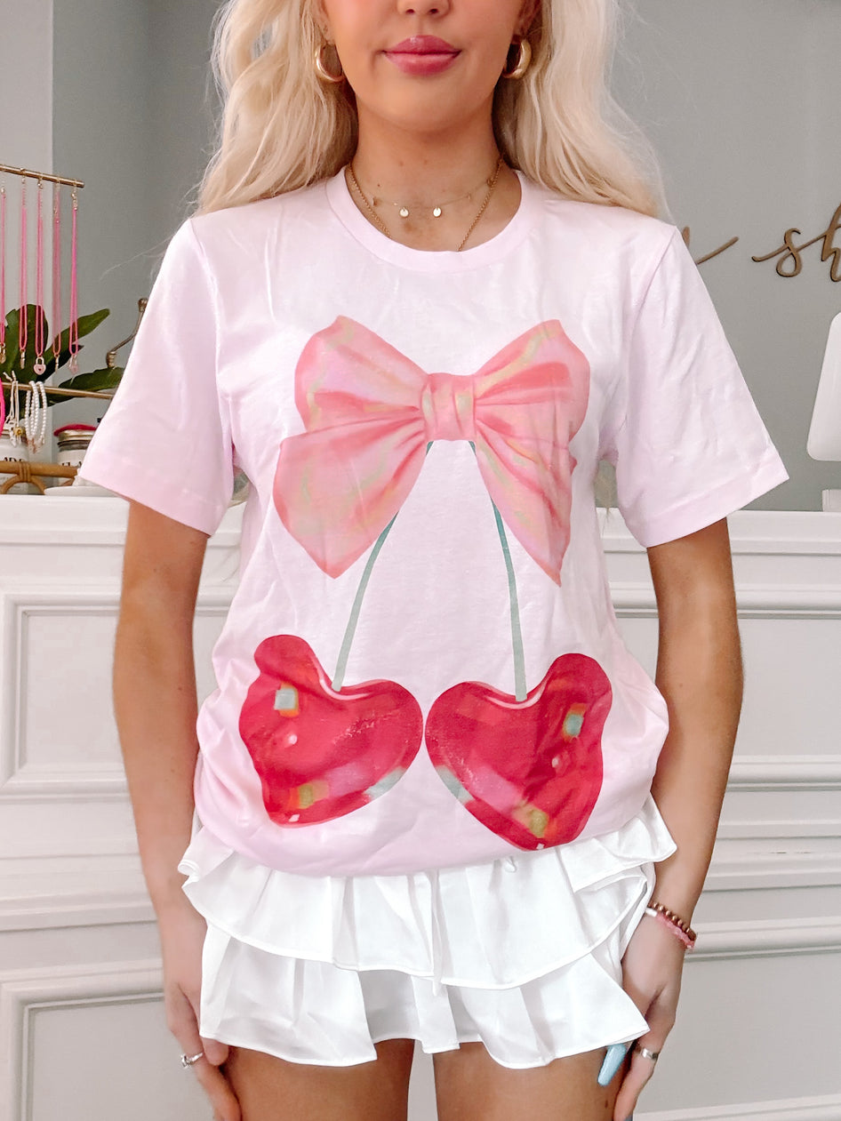 Pink t-shirt featuring a graphic of cherries with a pink bow, modeled on a person.
