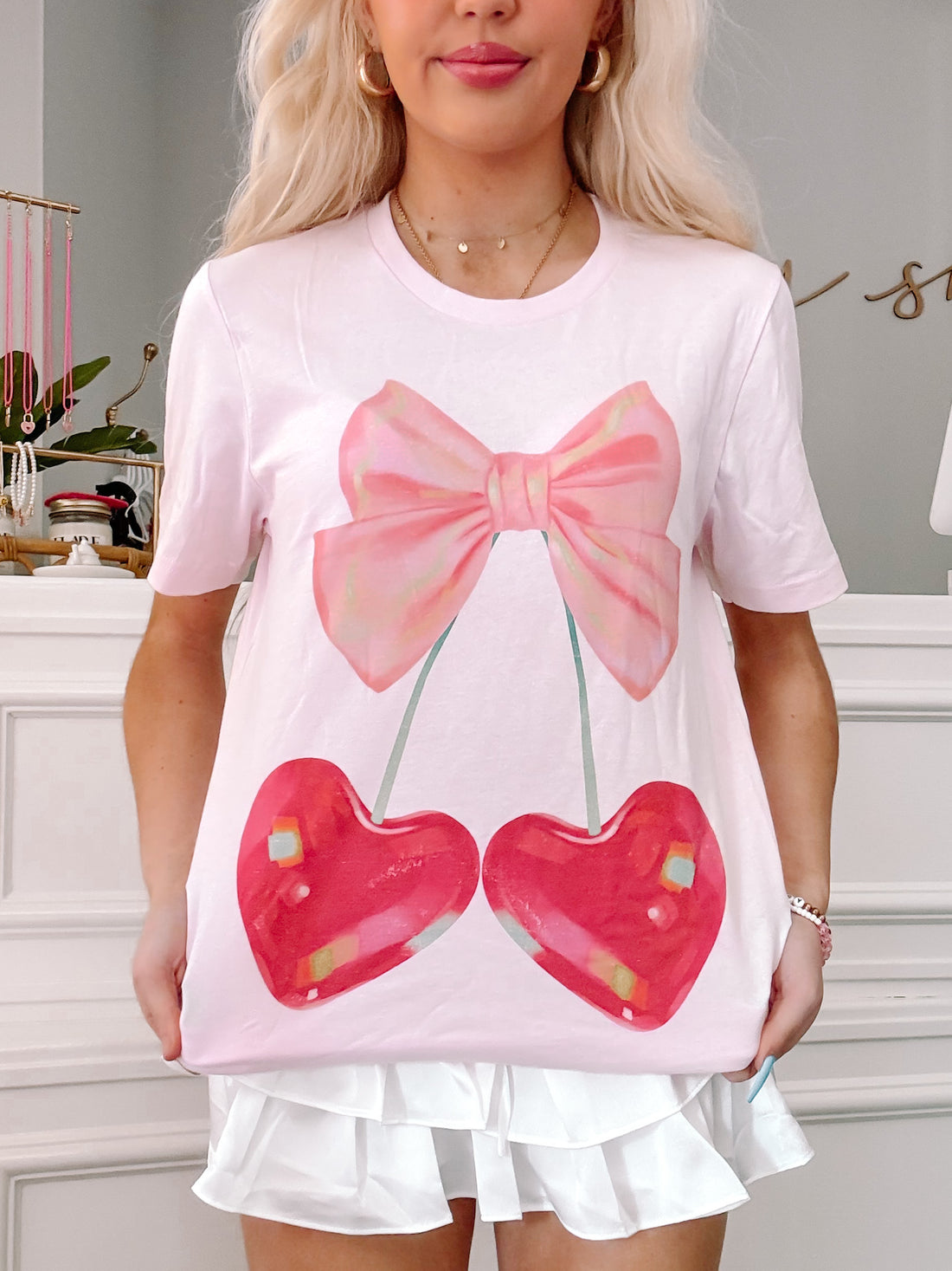 Pink t-shirt featuring a graphic of cherries with a pink bow, styled with a white pleated skirt.
