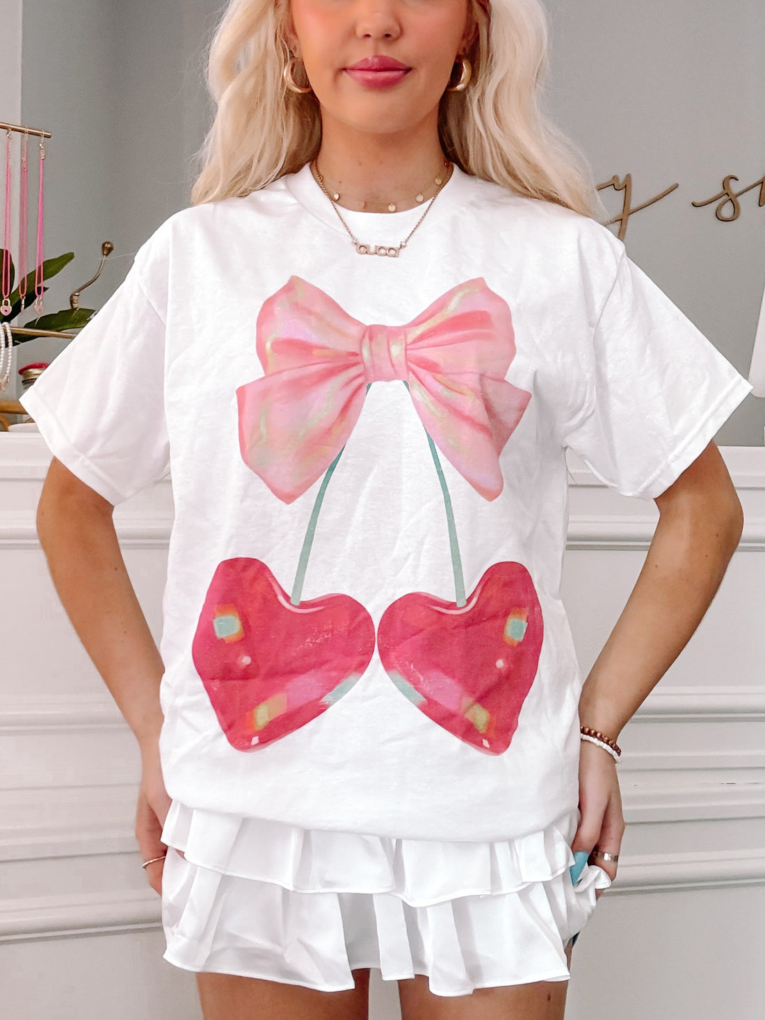 White tee showcasing a pink bow and two cherries; fun, summery graphic.
