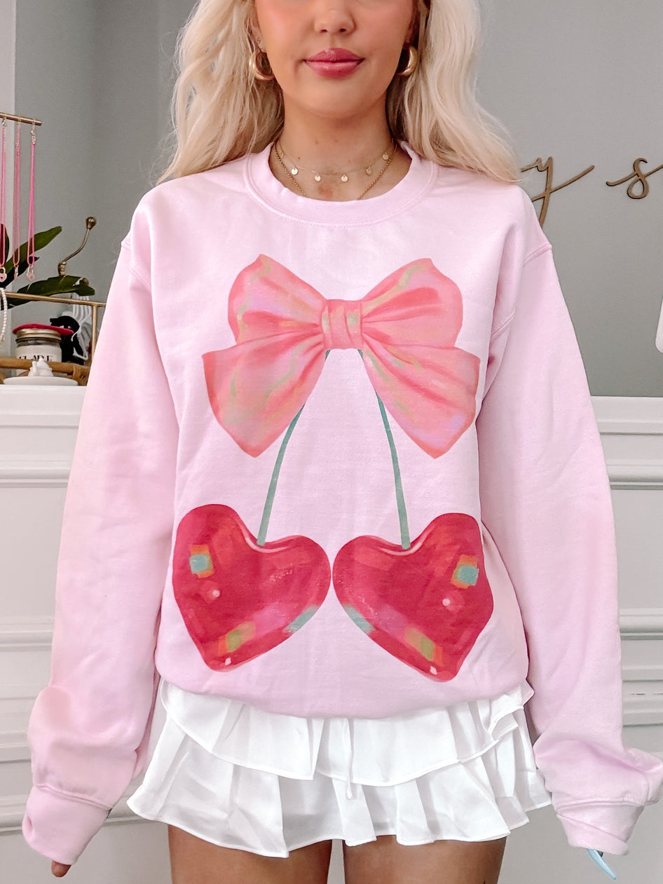 Pink crewneck sweatshirt featuring a whimsical illustration of cherries and a bow.
