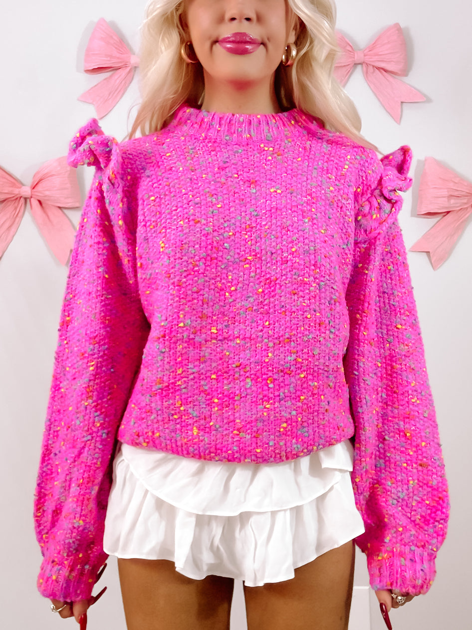Pink speckled knit sweater with ruffle details, modeled against a light background.
