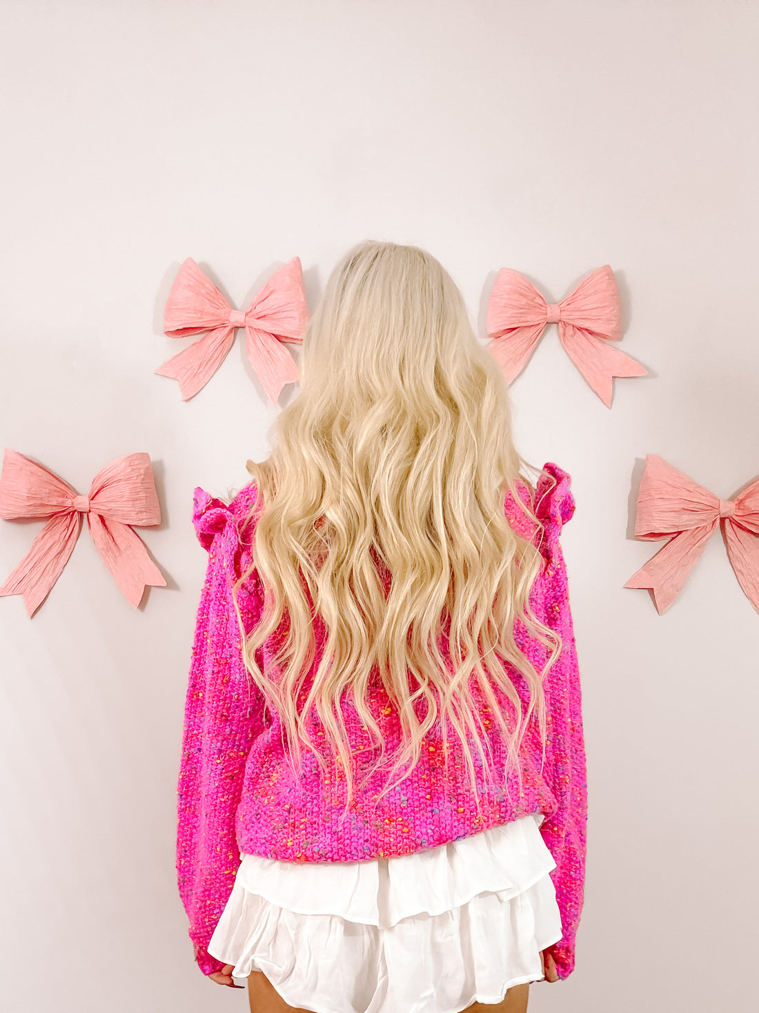 Pink confetti-pattern sweater styled with white tiered shorts, shown from the back against a wall decorated with pink bows.
