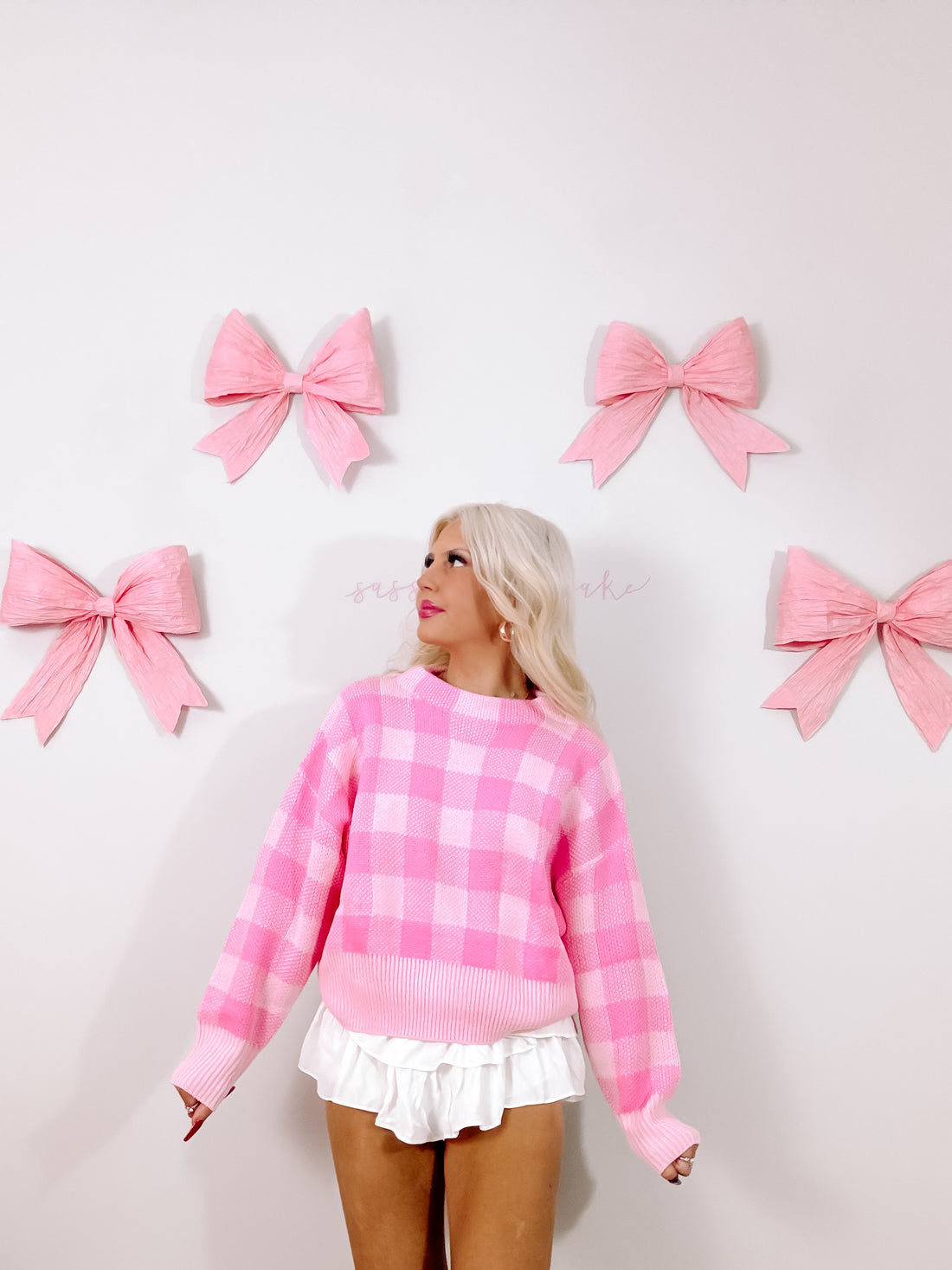 Pink gingham knit sweater styled against a wall adorned with pastel pink bows.
