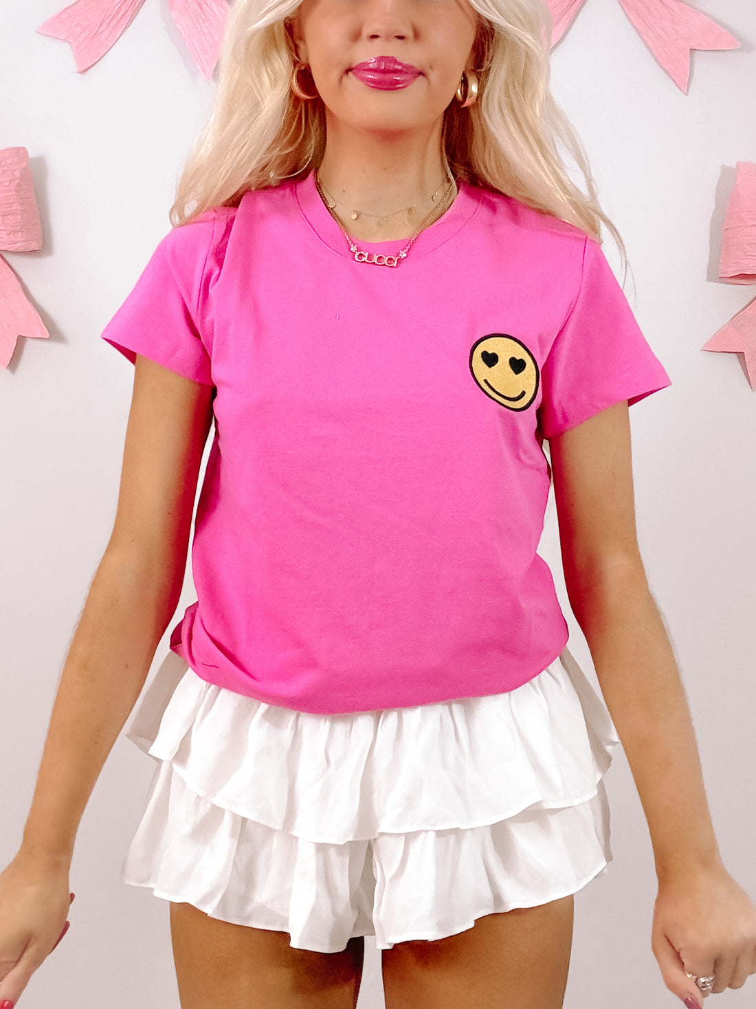 Pink short-sleeve tee with heart-eyed smiley face detail, styled with white ruffle shorts against a pink background.
