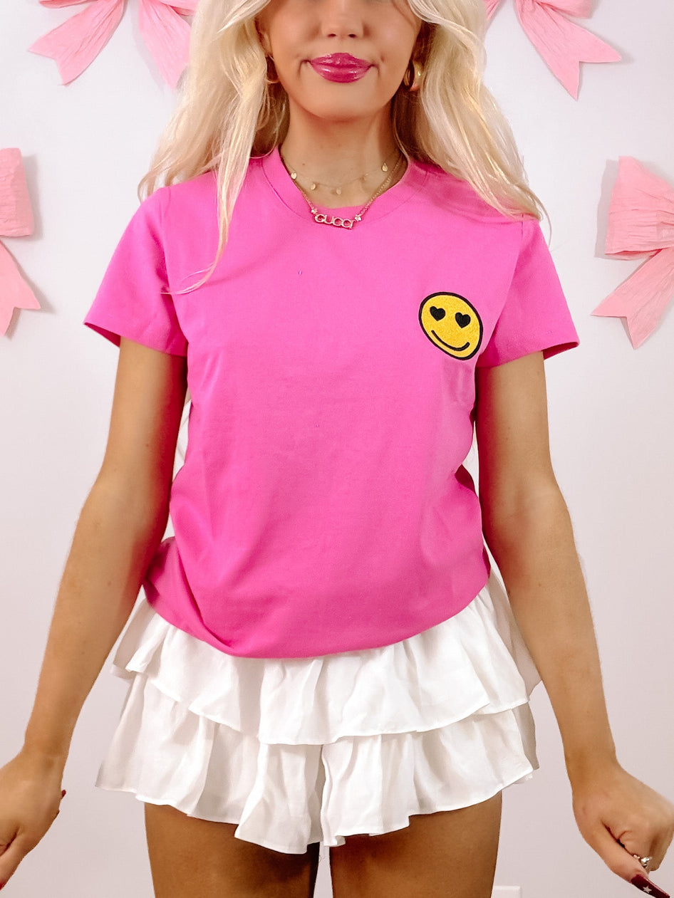 Hot pink short-sleeve tee with a cute heart-eyed smiley face graphic, modeled against a light background.
