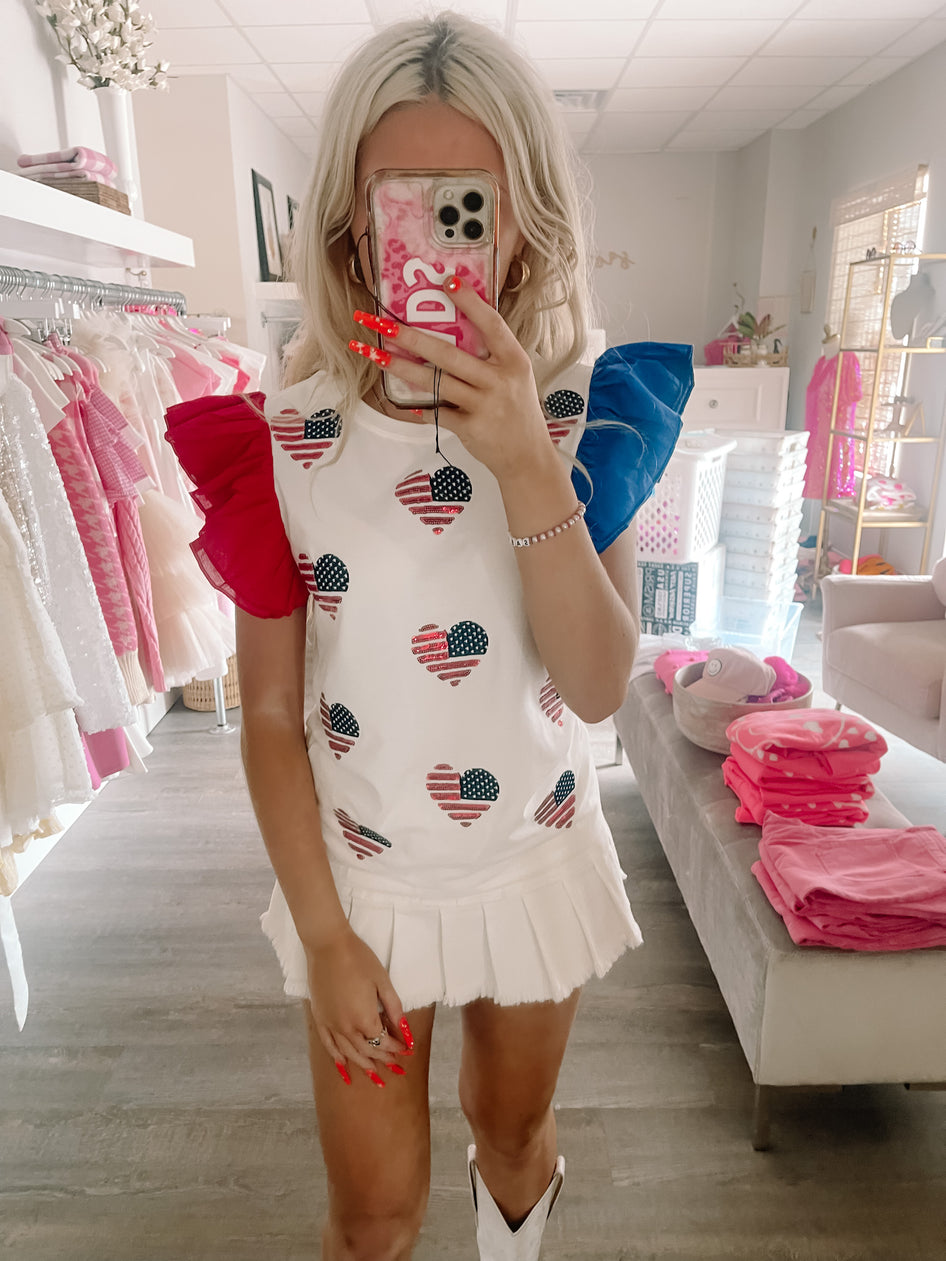 White ruffle top featuring American flag heart appliqués, shown on a model in a boutique setting.
