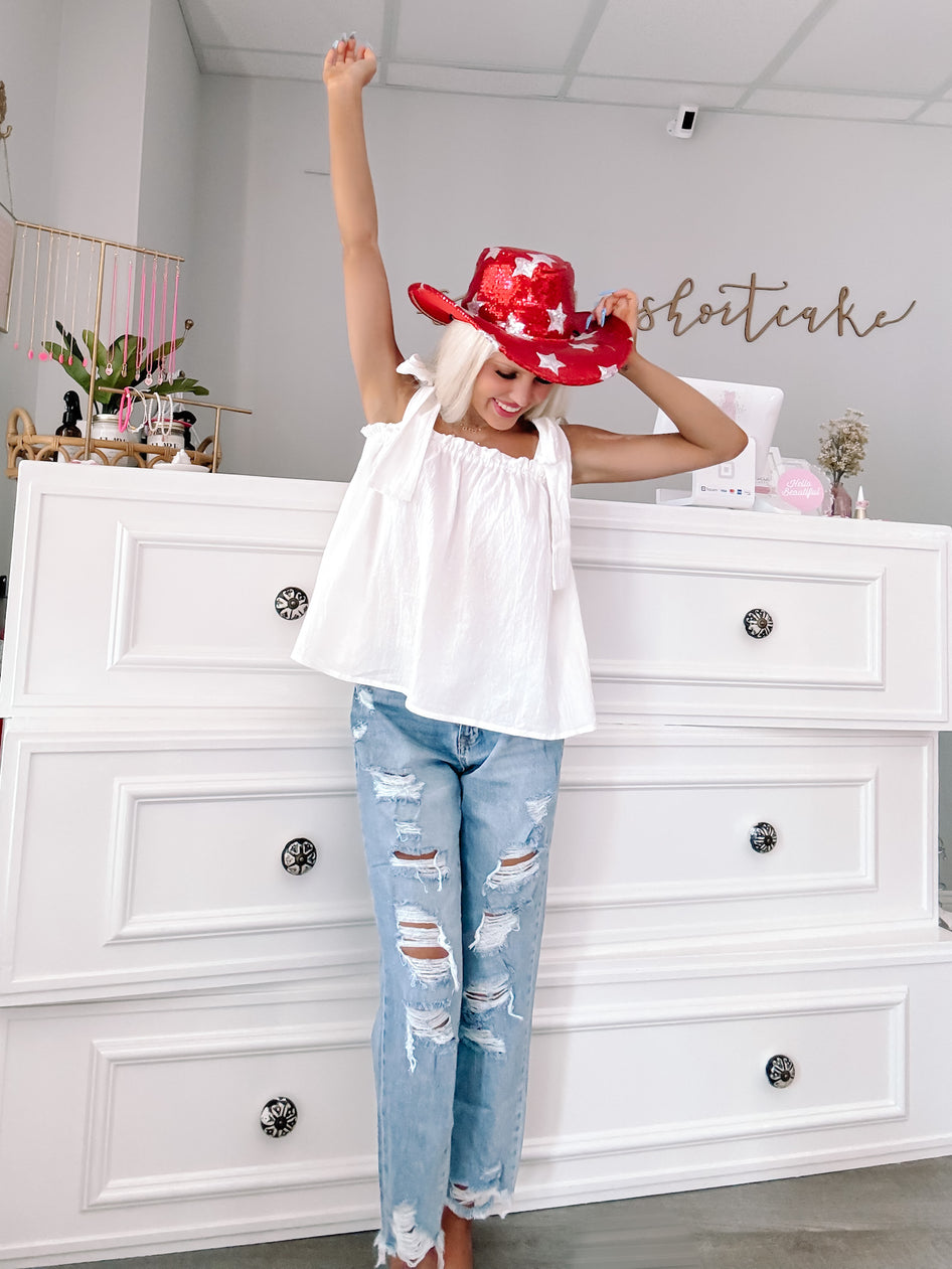 White eyelet ribbon top styled with distressed denim jeans and a red sequined cowboy hat.
