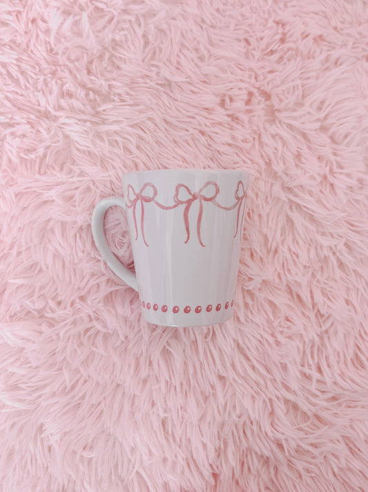 White ceramic mug with pink ribbon design, resting on a fluffy pink surface.