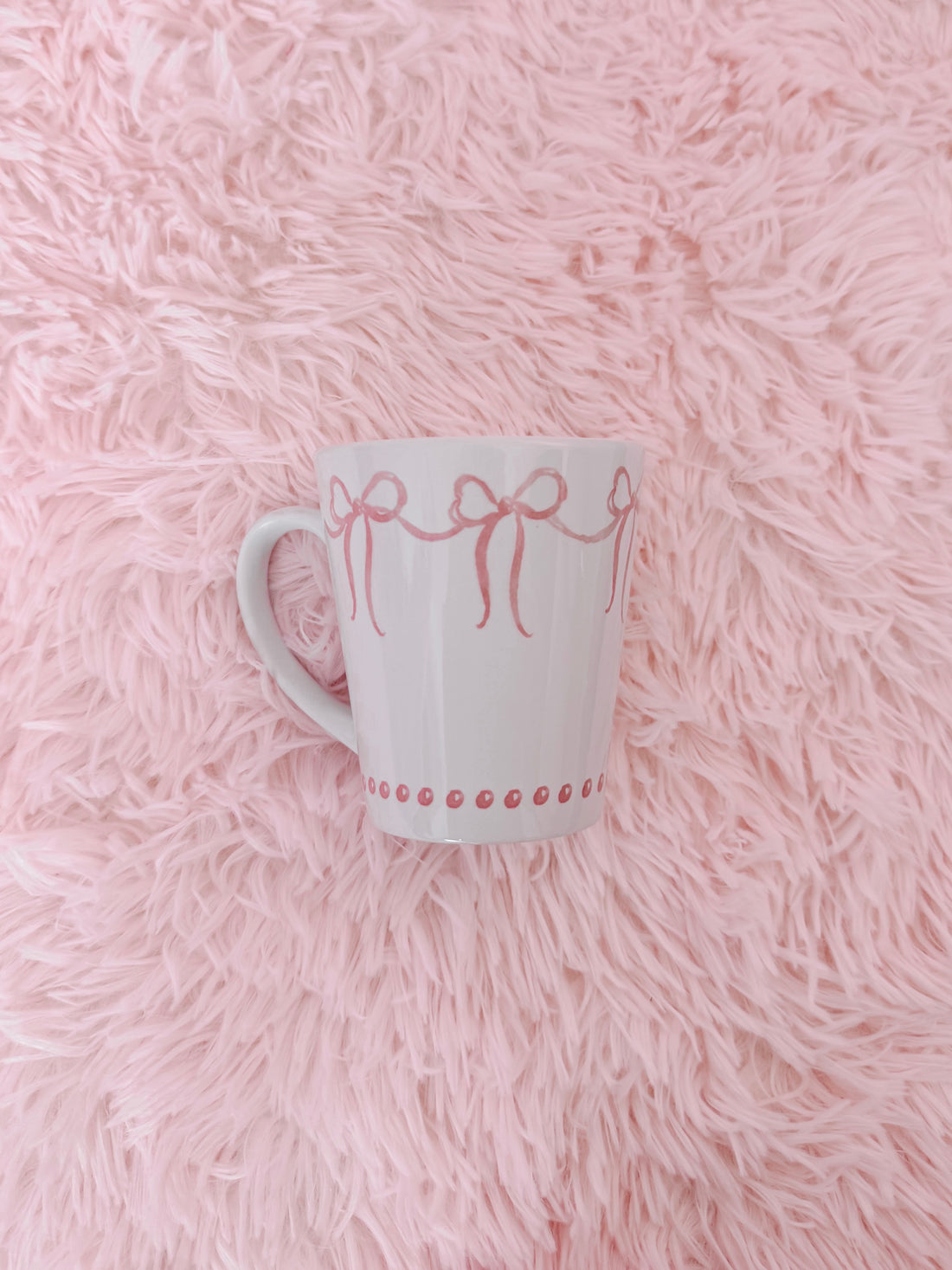White ceramic mug with pink ribbon design, resting on a fluffy pink surface.
