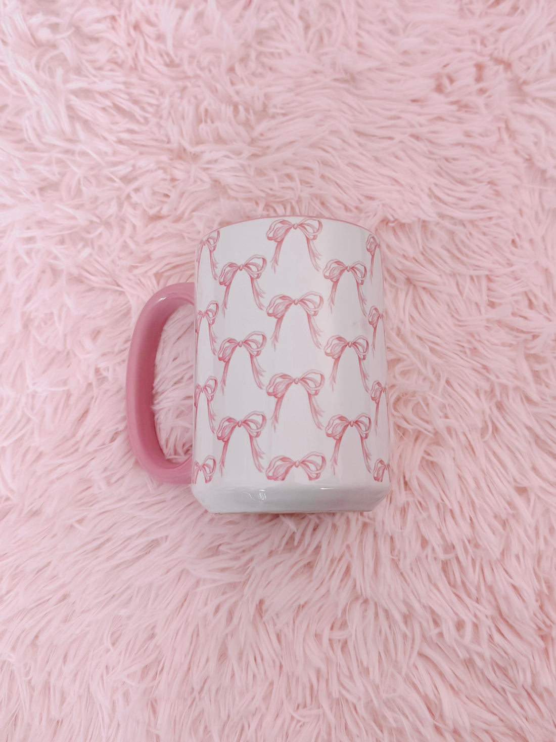 Pink ceramic mug with a delicate pink bow pattern, shown on a fluffy pink background.
