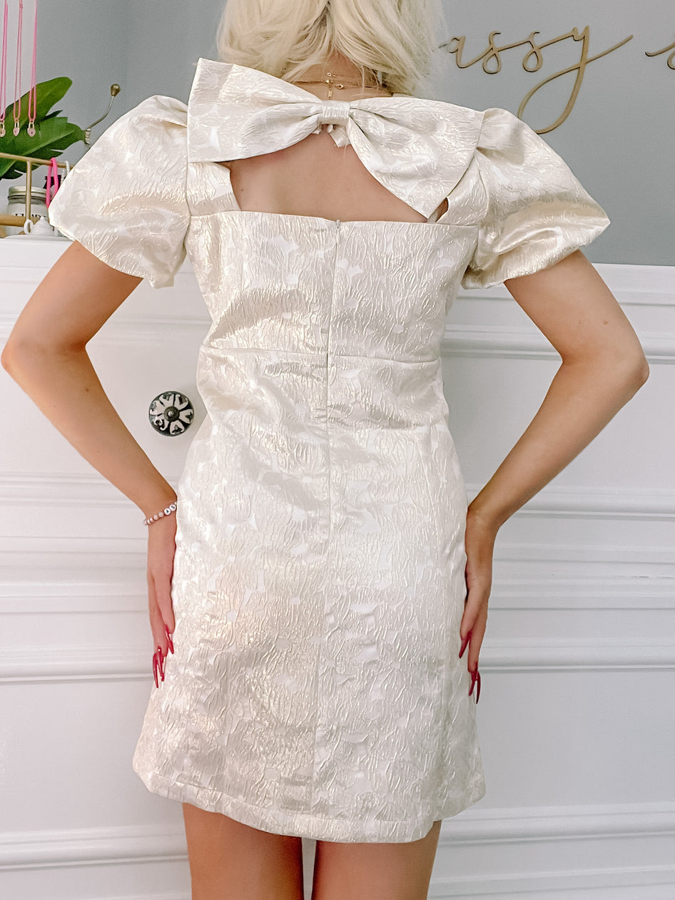 Gold mini dress with puff sleeves and a large bow detail at the back.
