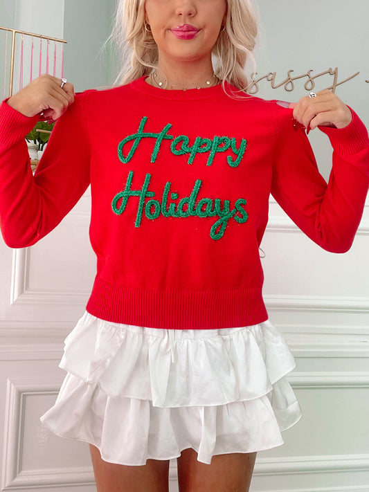 Red Christmas sweater with green sparkly "Happy Holidays" lettering, paired with a white ruffled skirt.
