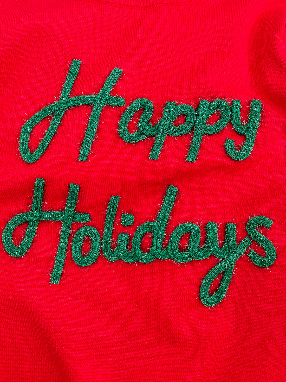 Red knit sweater detail featuring green embroidered "Happy Holidays" lettering.

