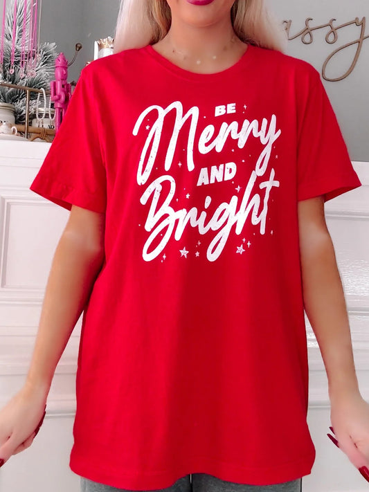 Person wearing a red t-shirt with 'Be Merry and Bright' text in a festive setting.