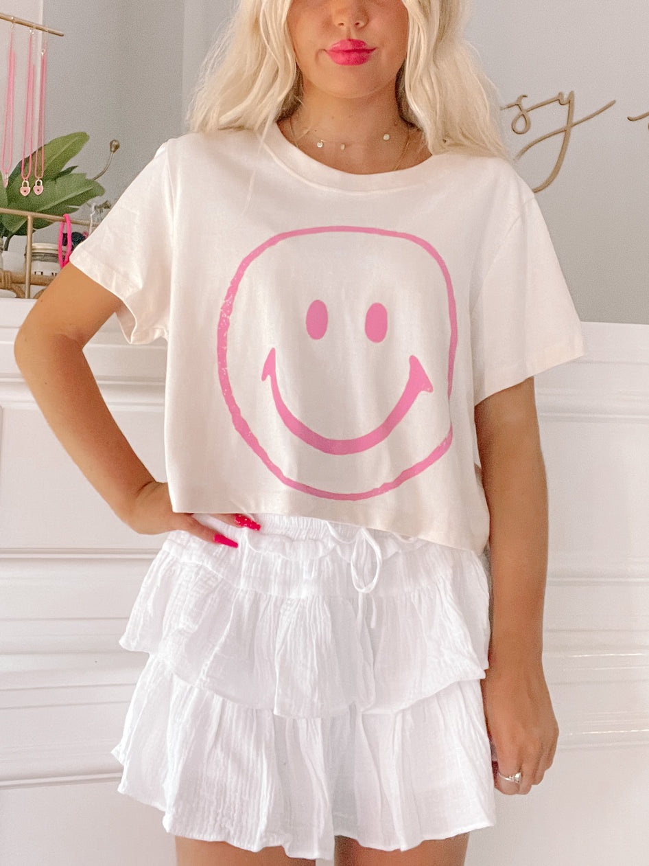 Cream cropped tee featuring a pink smiley face graphic, styled with a white tiered skirt.

