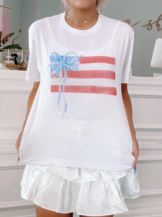 White tee featuring a watercolor-style American flag design with a blue bow; casual summer style.