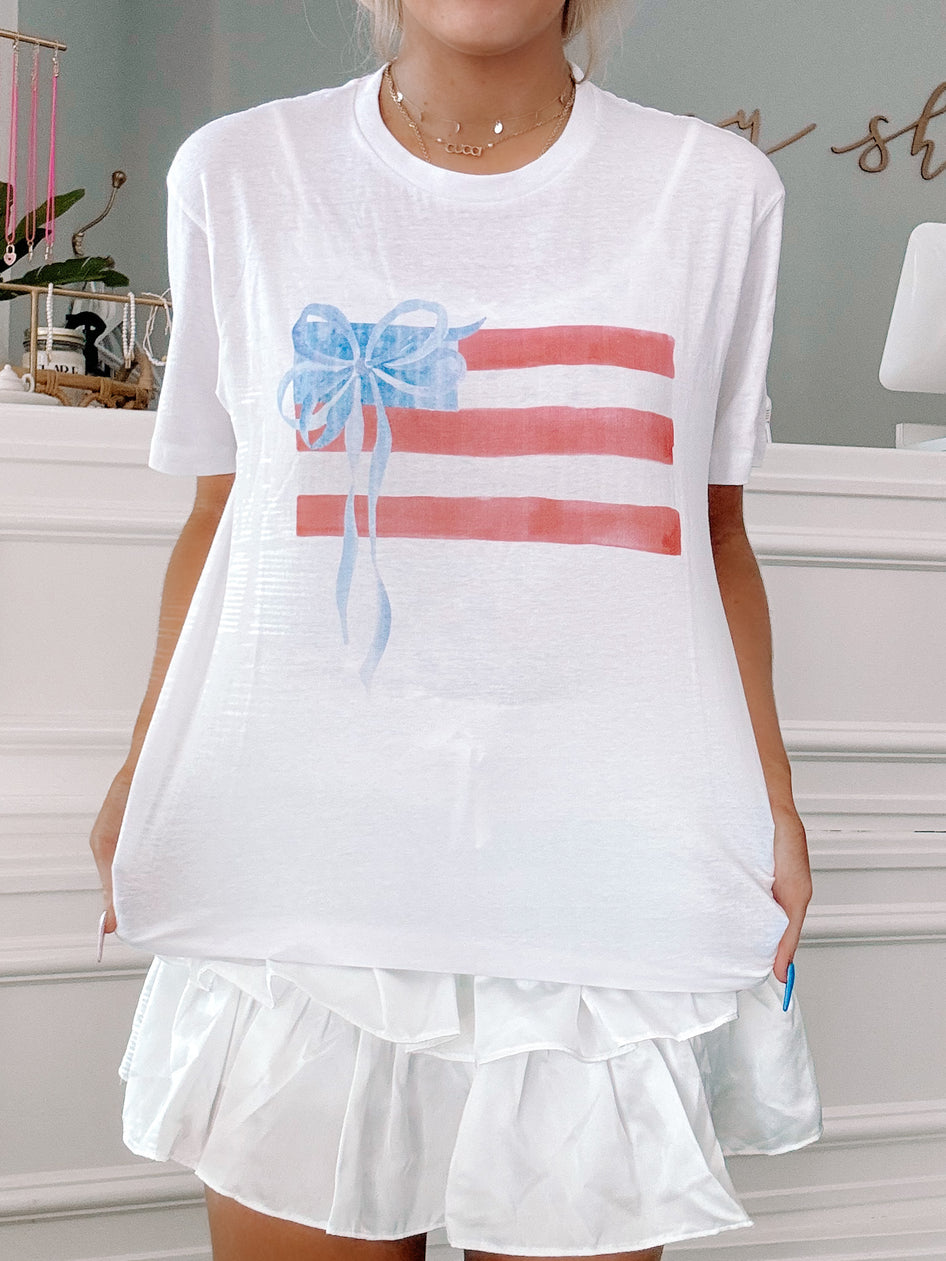 White tee featuring a watercolor-style American flag design with a blue bow; casual summer style.
