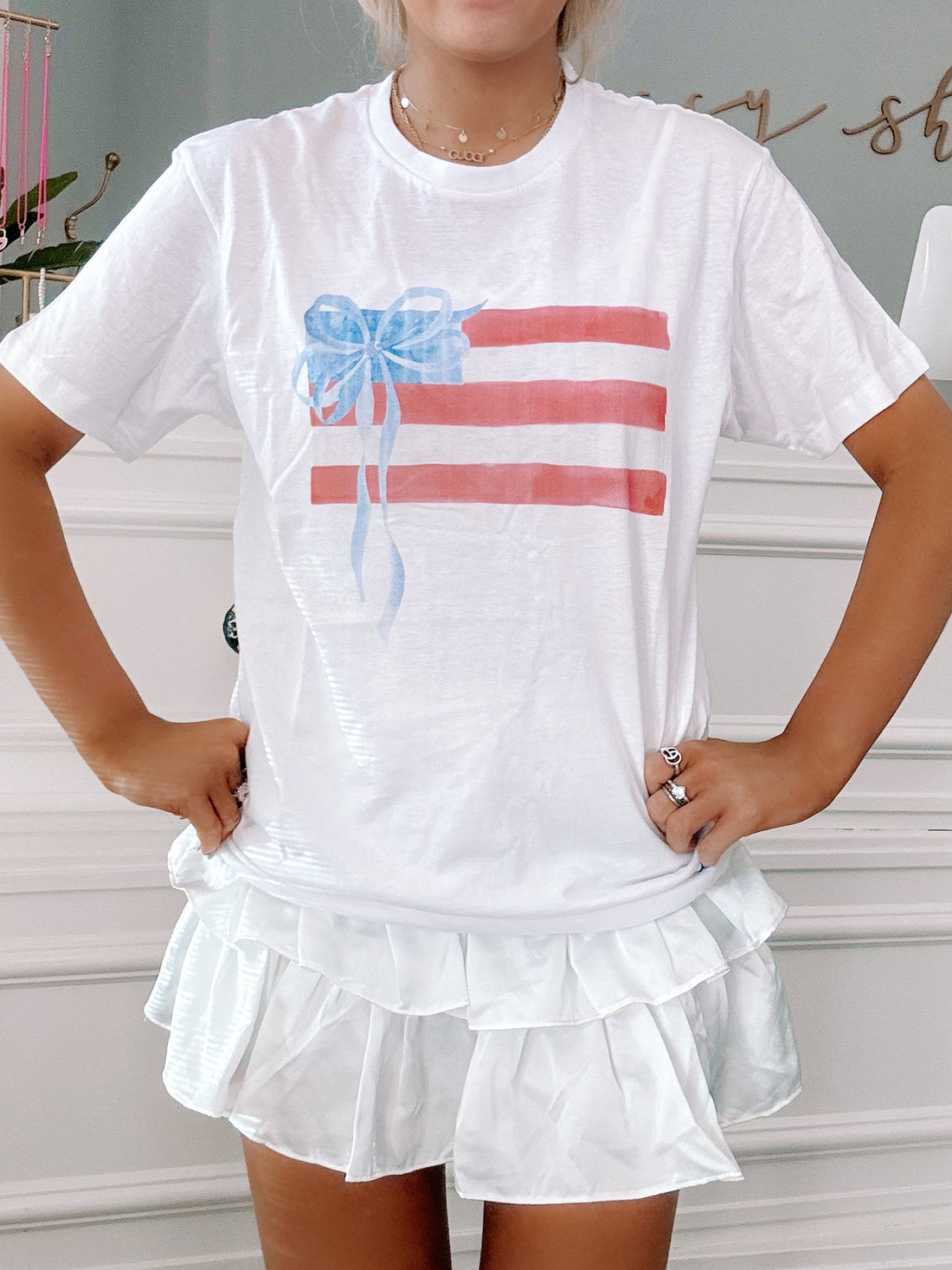 White tee featuring a watercolor-style American flag design with a blue bow, modeled on a person.
