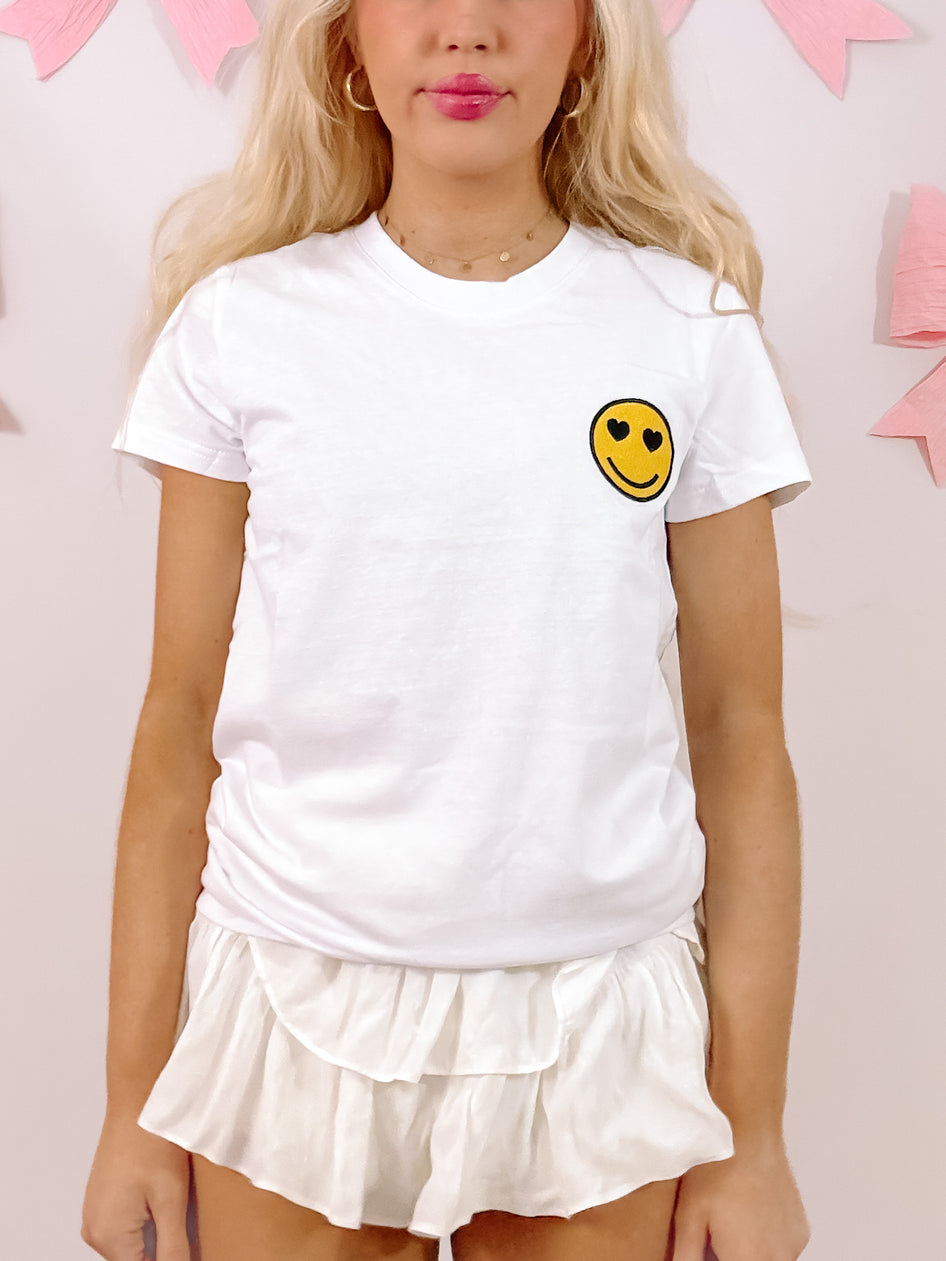 White Happy Heart Tee featuring a yellow embroidered smiley face with heart eyes.
