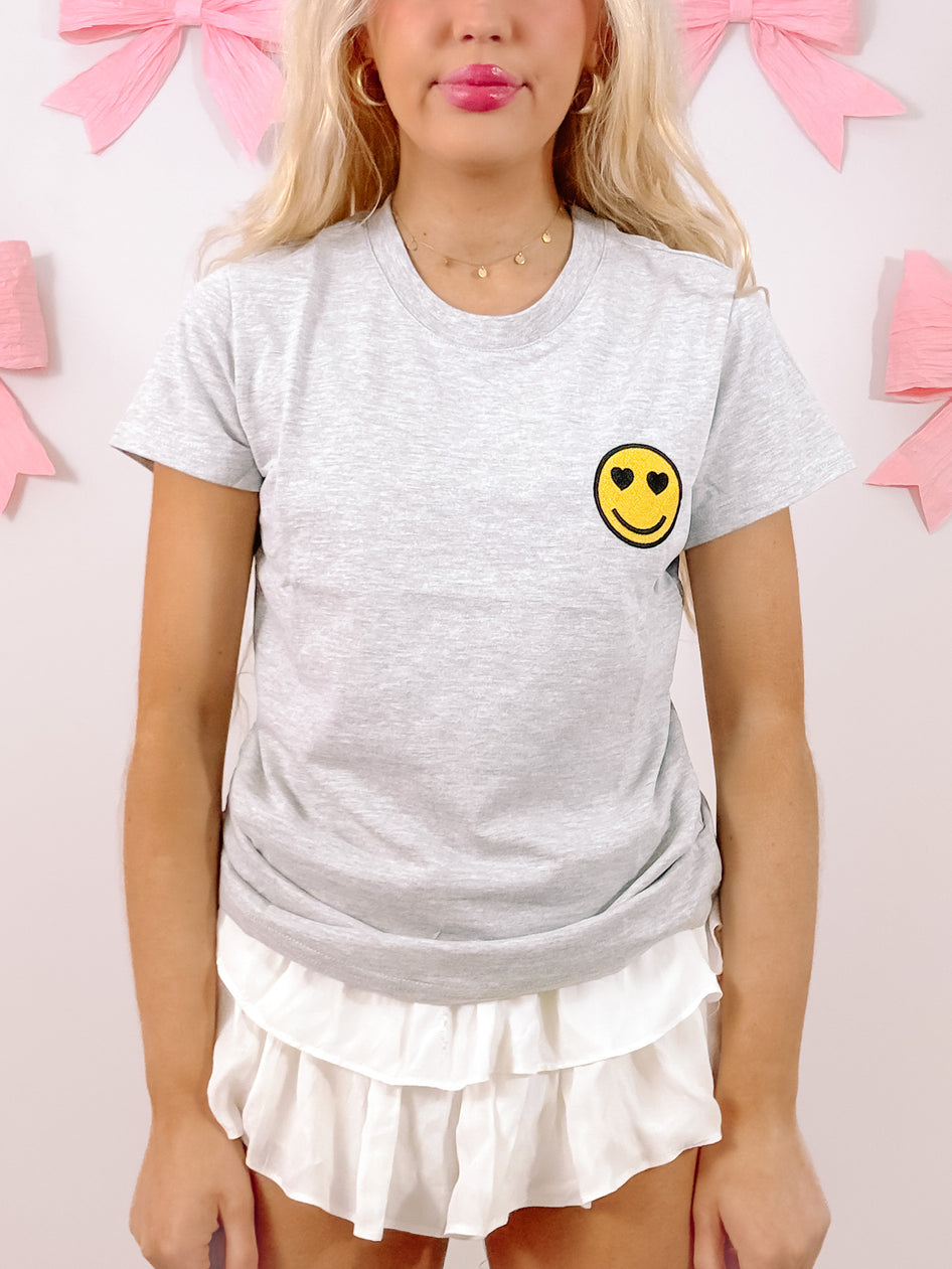 Grey short-sleeve tee featuring a small, embroidered smiley face with heart eyes;  model shown wearing with white ruffled shorts.
