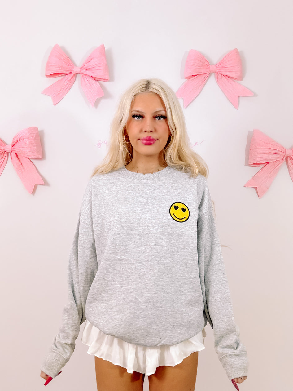 Grey crewneck sweatshirt featuring a small, heart-eyed smiley face detail; styled against a pink backdrop.
