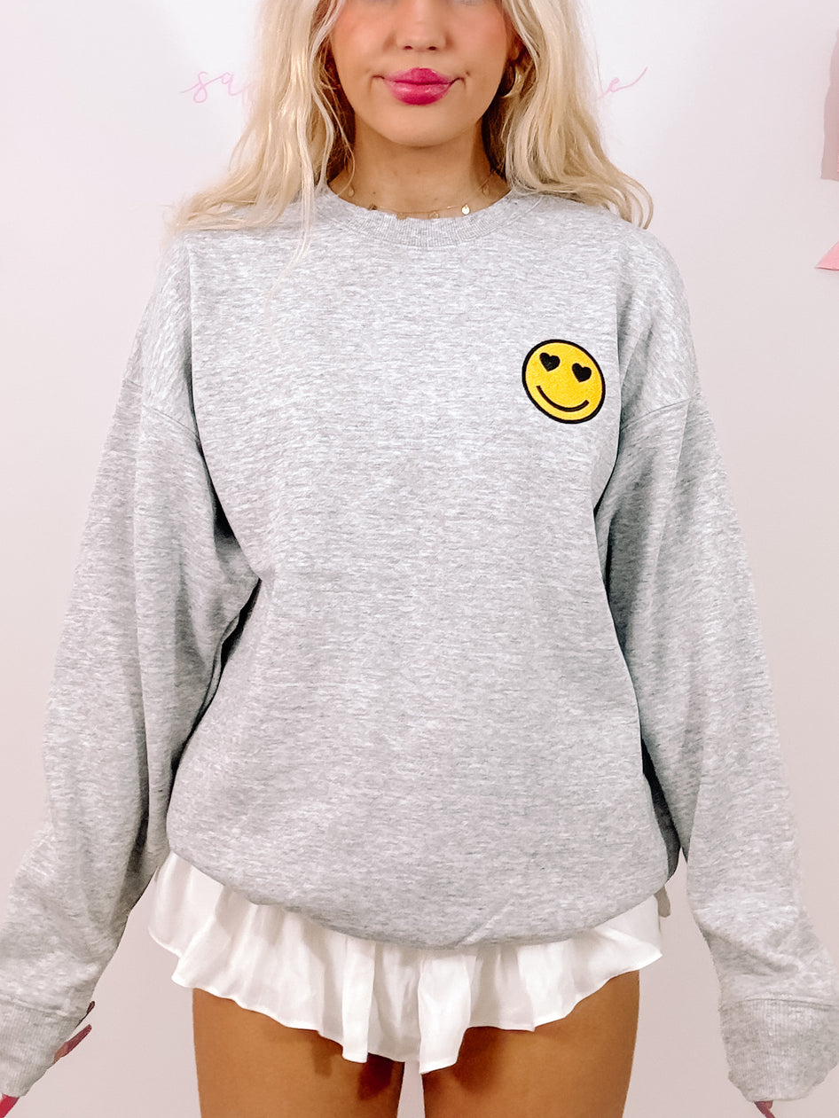 Grey crewneck sweatshirt with a small, yellow smiley face emblem featuring heart-shaped eyes.
