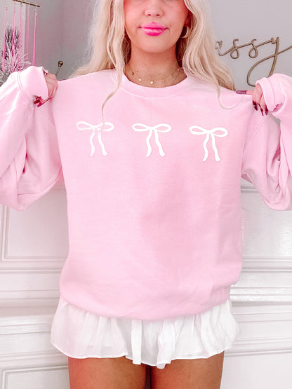 Pink crewneck sweatshirt featuring three white bows, modeled against a white wall.
