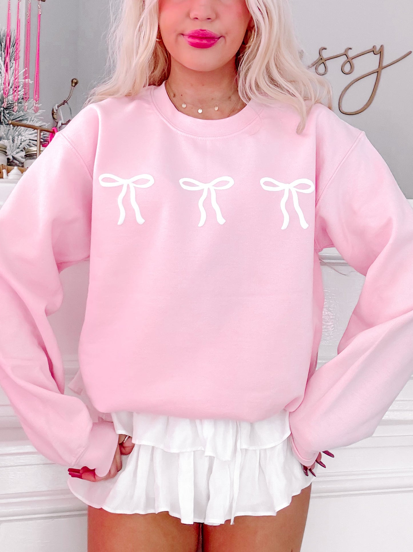 Pink crewneck sweatshirt featuring three white bows, modeled against a neutral backdrop.
