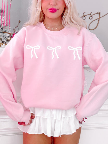 Pink crewneck sweatshirt featuring three white bows, modeled against a neutral backdrop.
