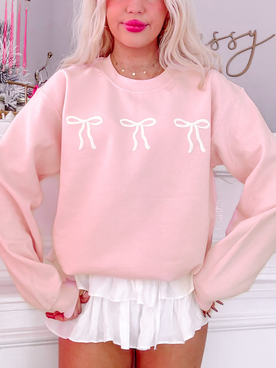 Pink crewneck sweatshirt featuring three embroidered bows;  model wears it with a white skirt.
