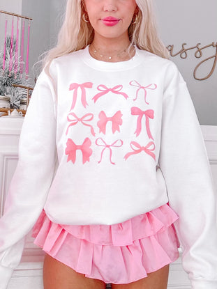 White crewneck sweatshirt featuring a pink bow print, modeled with a pink ruffled skirt.

