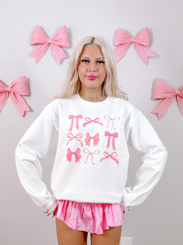 White crewneck sweatshirt featuring a pink bow print, modeled against a wall decorated with large pink bows.
