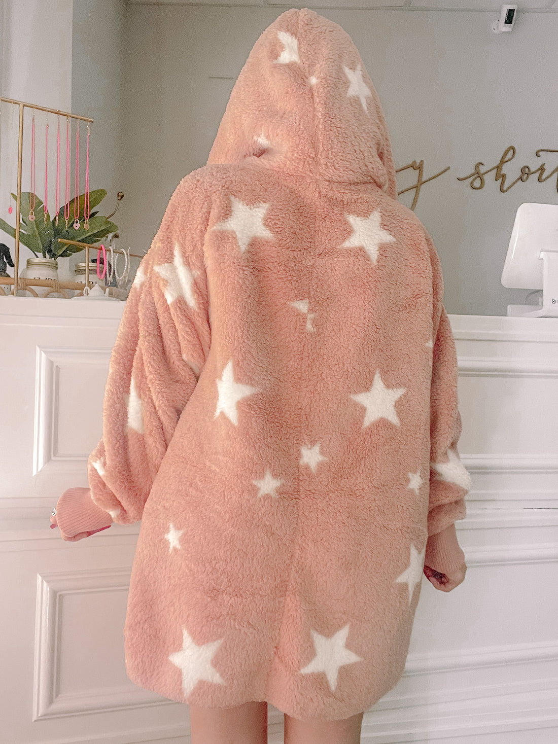 Pink fuzzy hooded jacket adorned with white stars; cozy sweater shown from behind in a shop setting.
