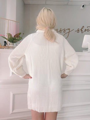 Cream-colored ribbed knit sweater dress, shown from behind in a boutique setting.
