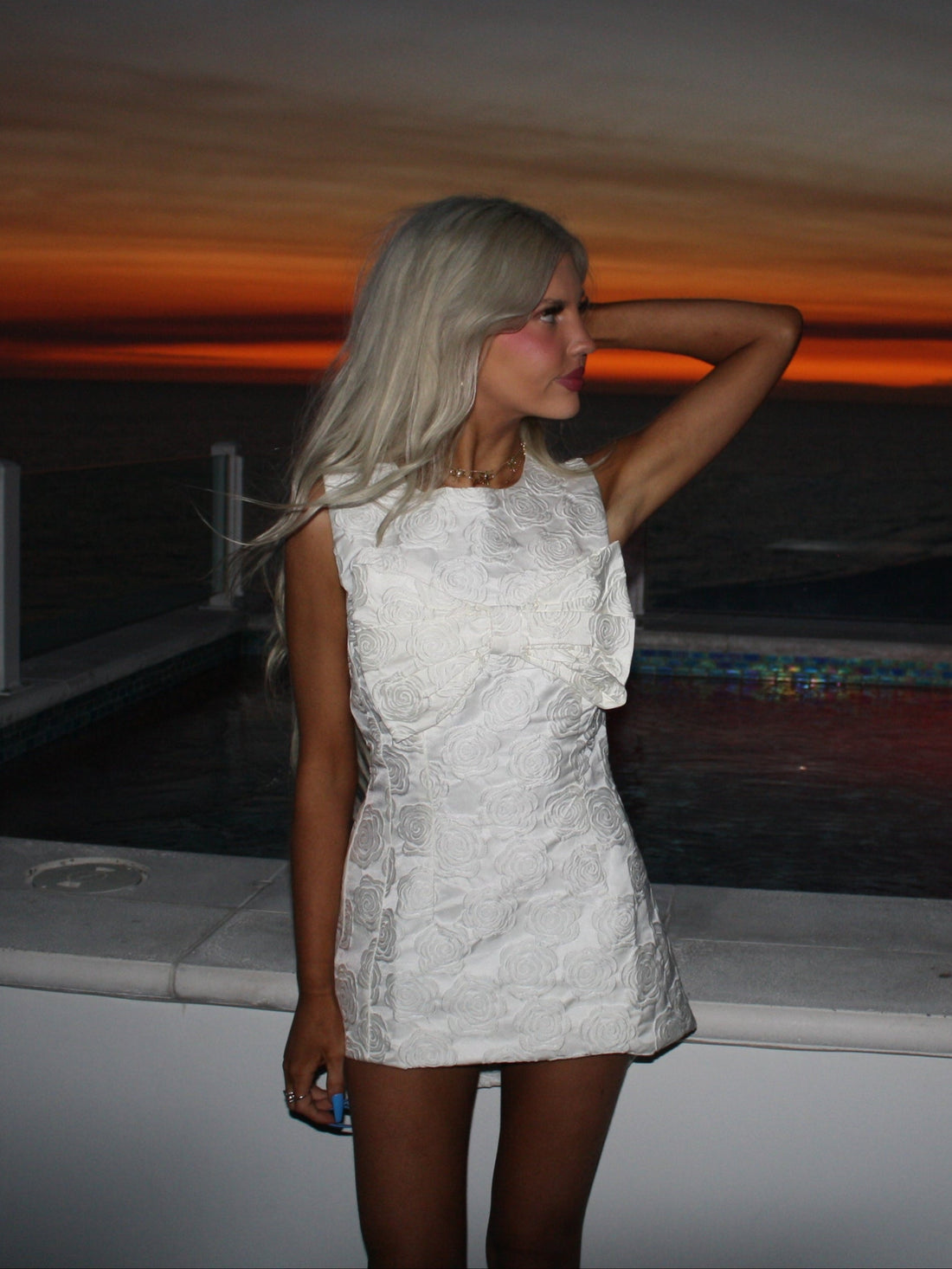 White rose-patterned mini dress, worn by a model at sunset by a pool.
