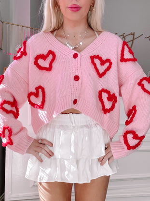 Pink knit cardigan adorned with red heart appliqués, modeled over a white tiered mini skirt.
