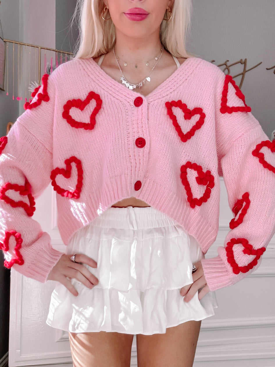Pink knit cardigan adorned with red heart appliqués, modeled over a white tiered mini skirt.
