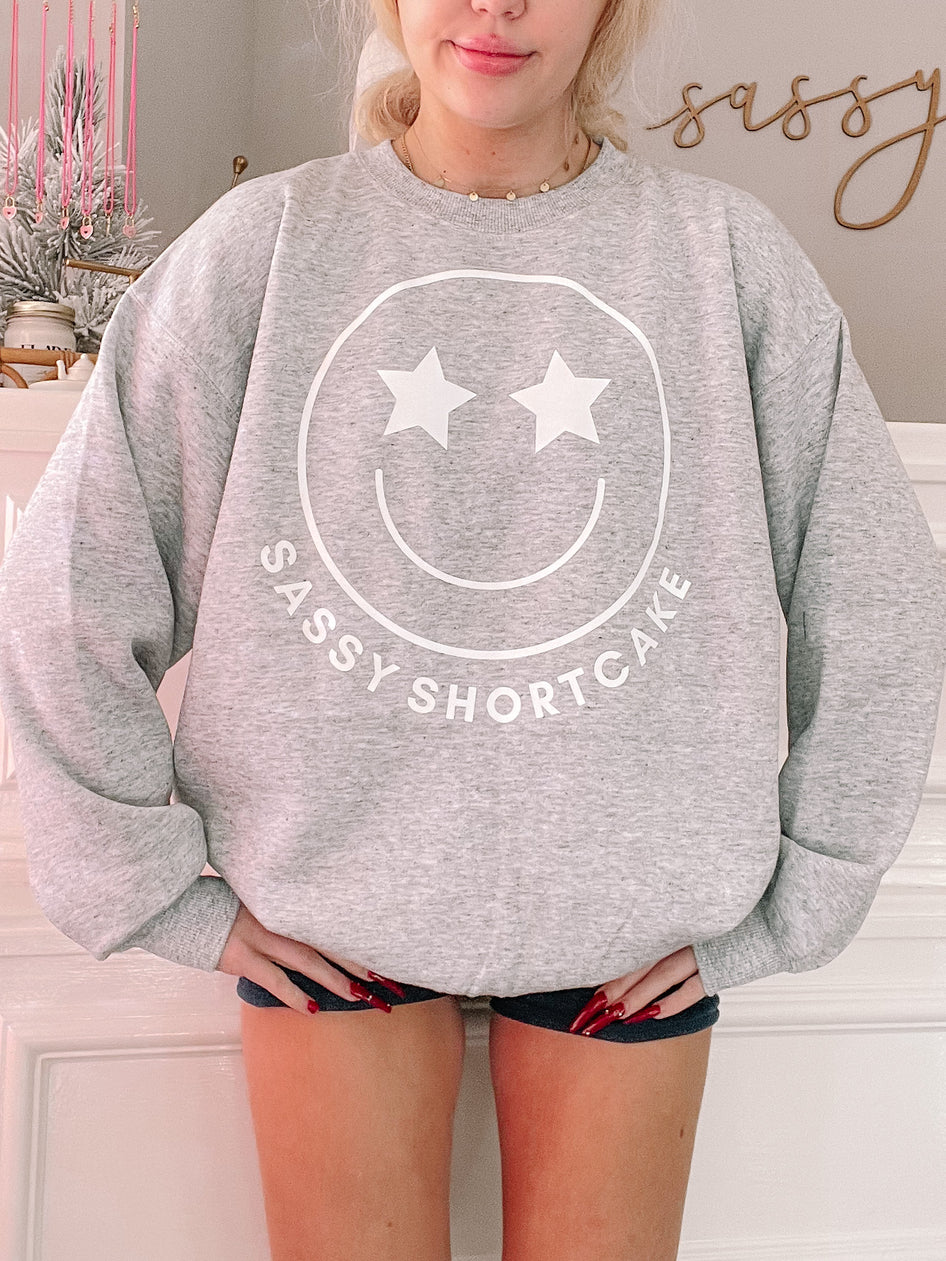 Grey Sassy Shortcake Smiley Crewneck sweatshirt with star-eye smiley face graphic.
