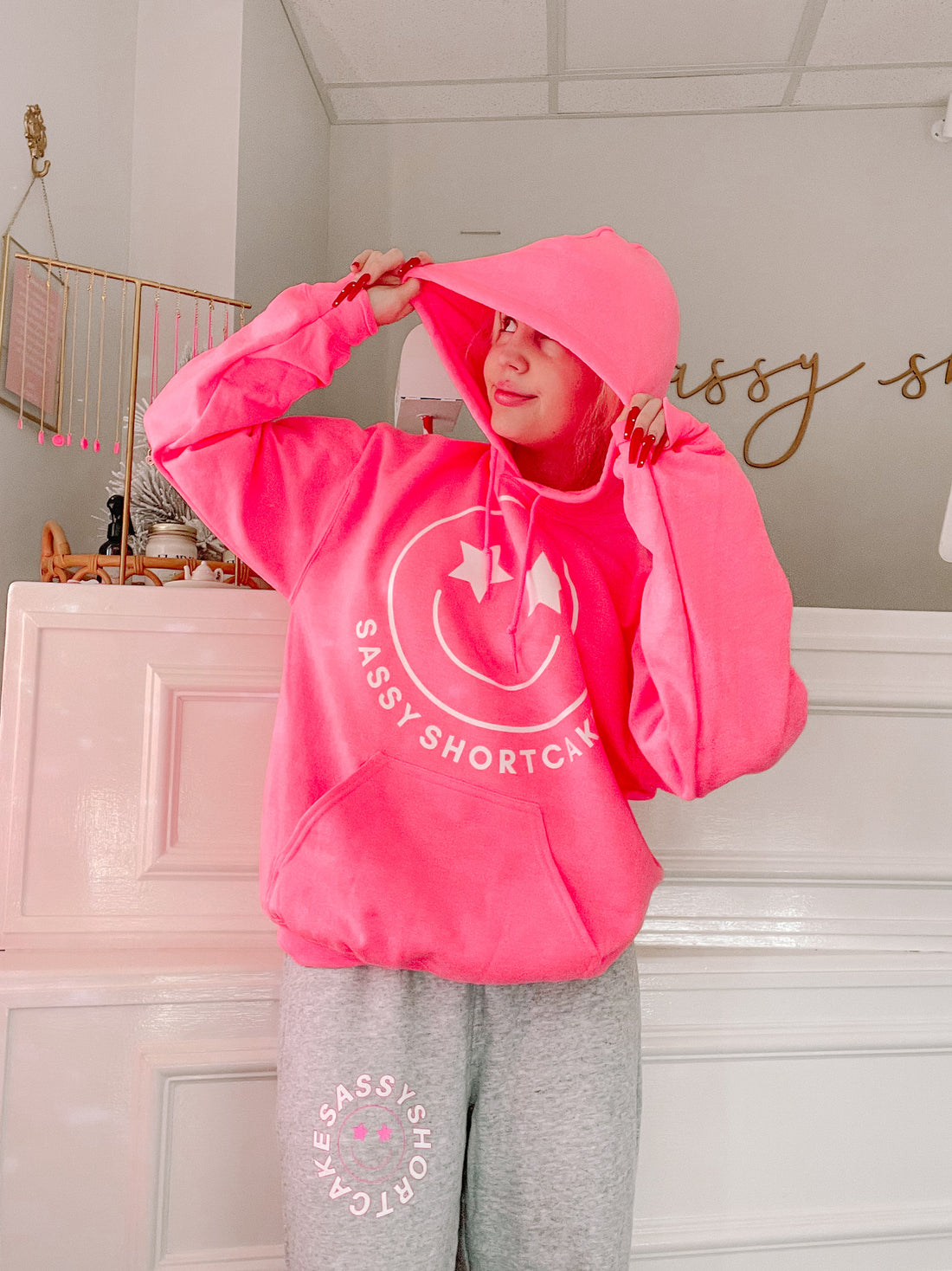 Bright pink hoodie featuring a smiley face design, modeled against a white paneled wall.
