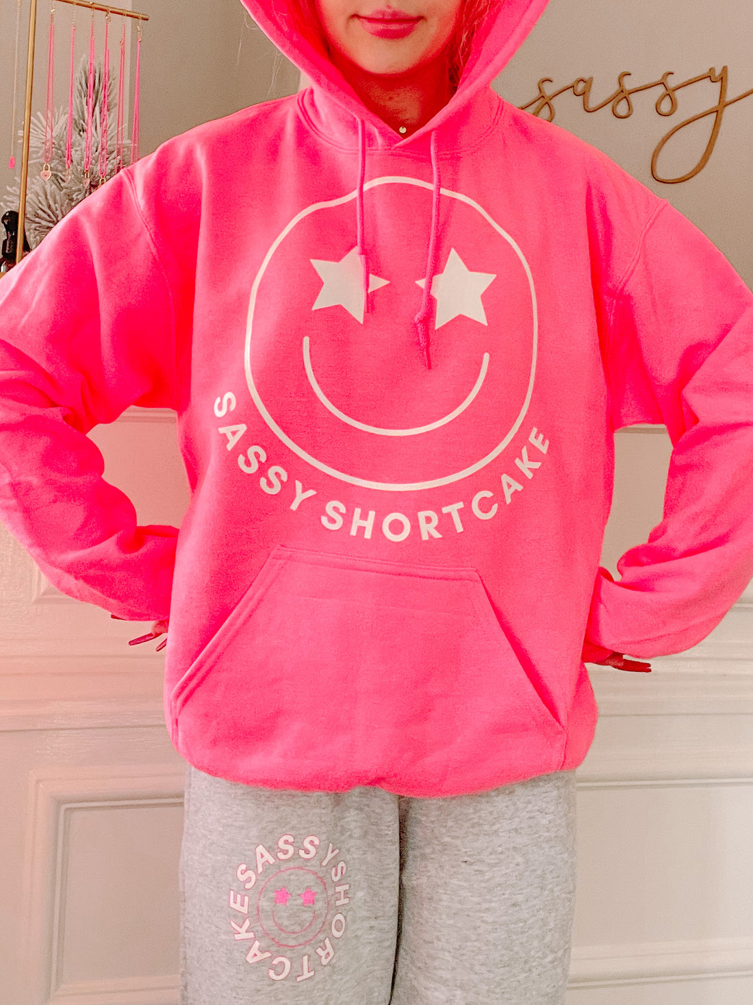 Bright pink hoodie featuring a smiley face graphic with stars for eyes, shown on a model.
