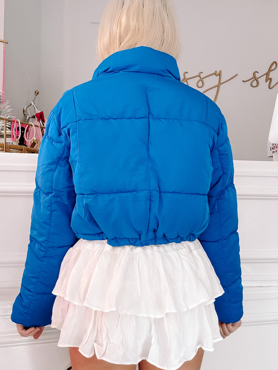 Bright blue puffer jacket, cropped style, shown from the back with a white ruffled skirt.
