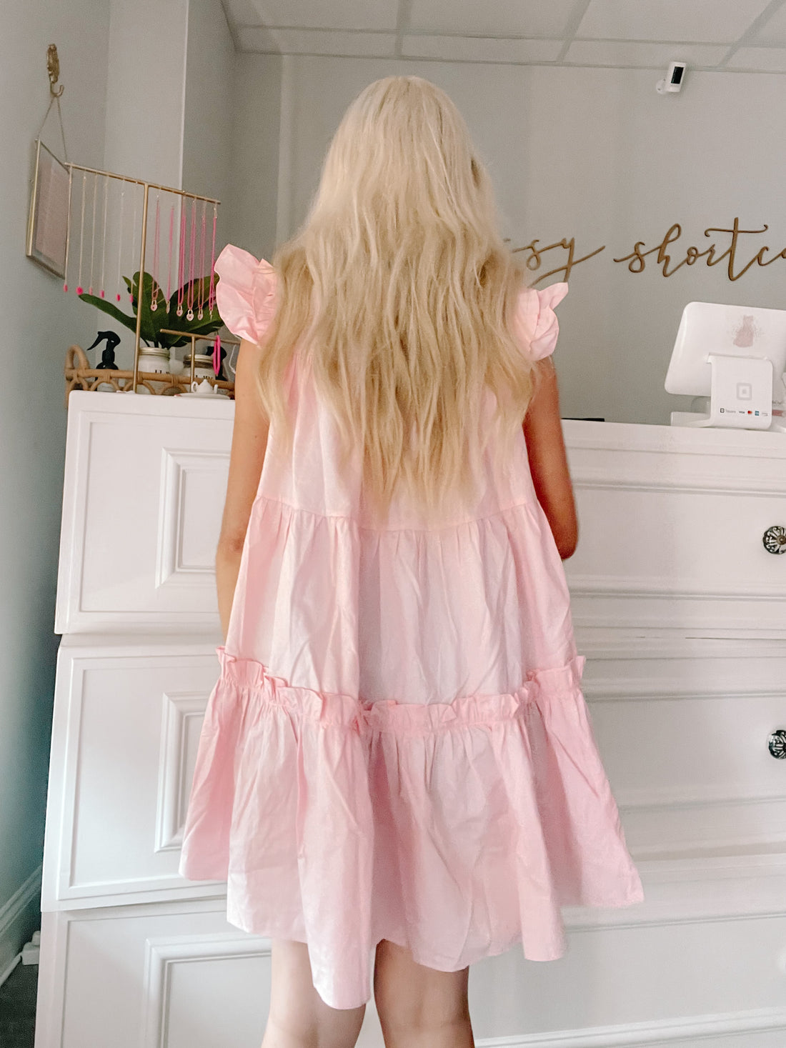Pink tiered mini dress with ruffle details, shown from the back in a boutique setting.
