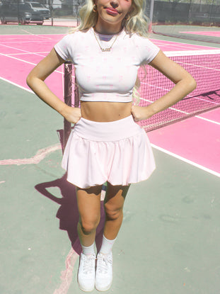 Model wearing white ribbed crop top and bubble skirt on pink tennis court.
