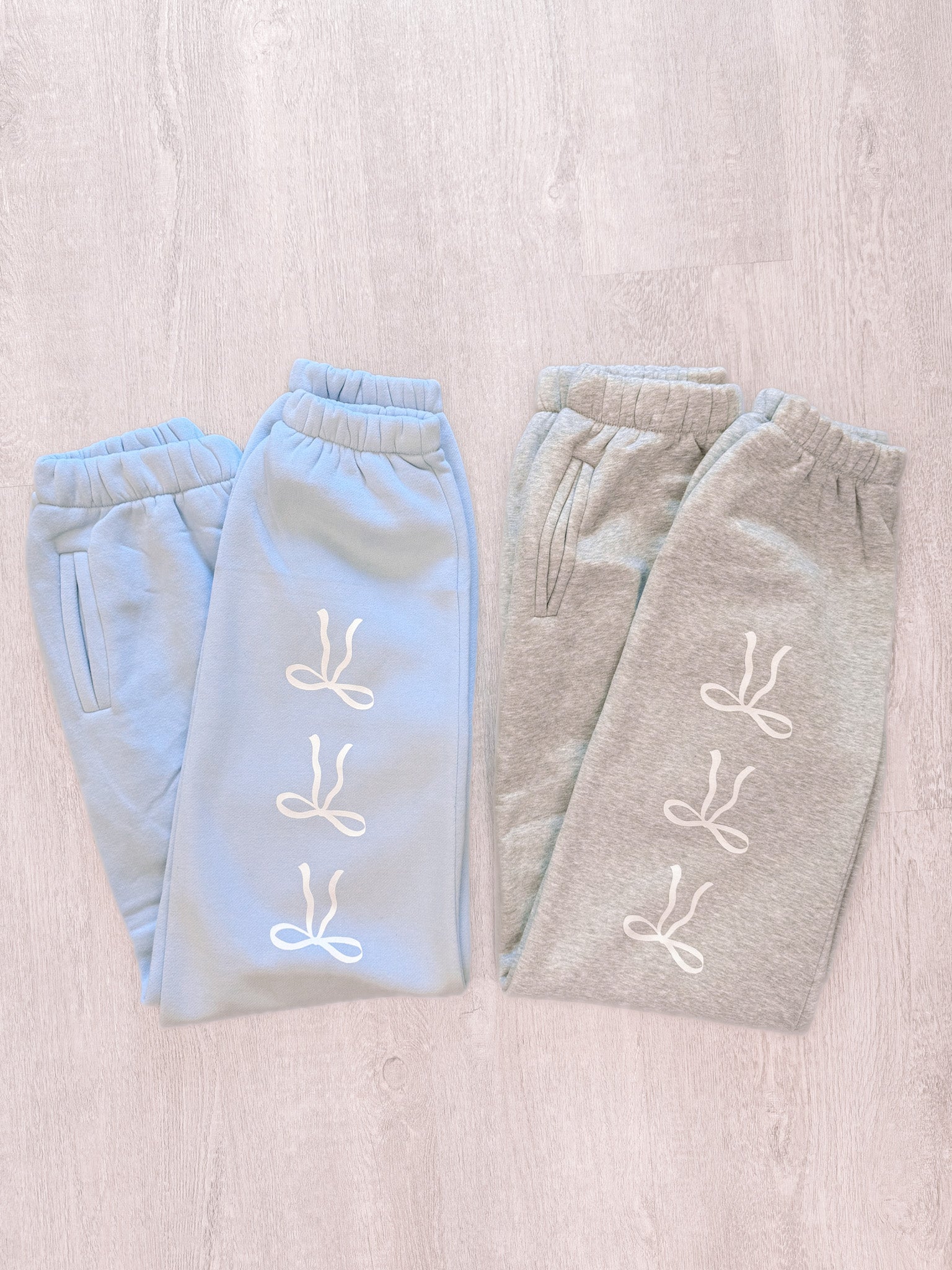 Light blue and gray sweatpants with a repeating bow design, folded on a wood background.
