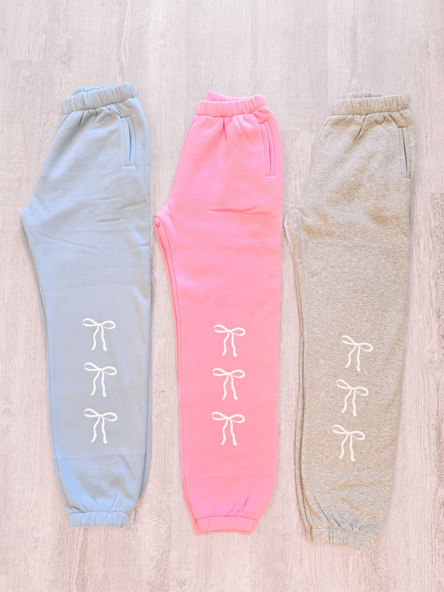 Light blue, pink, and gray sweatpants with white bow details, displayed on a wood background.
